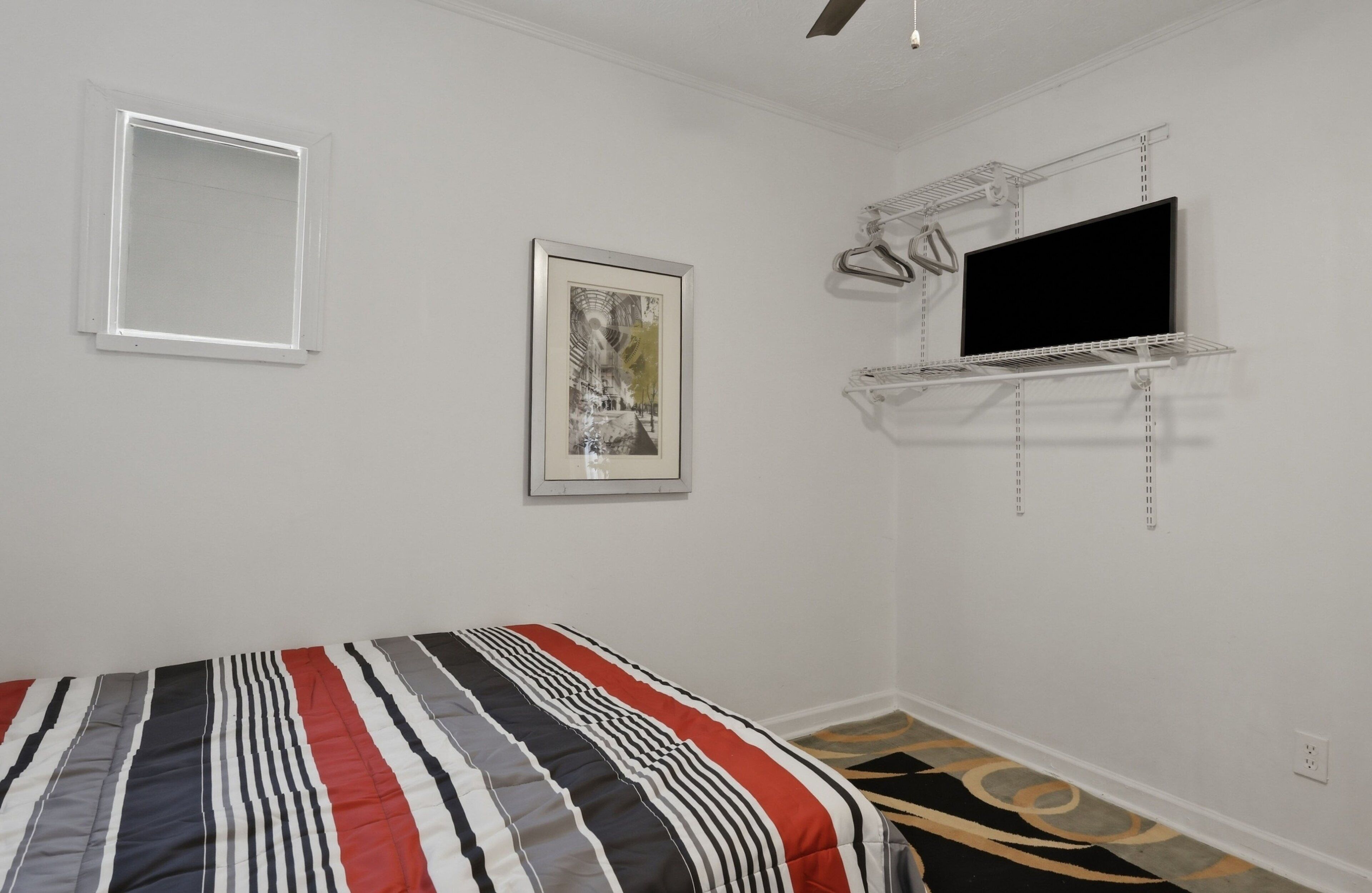 A bedroom with a bed, a framed picture, a TV on a wall-mounted shelf, and a window.