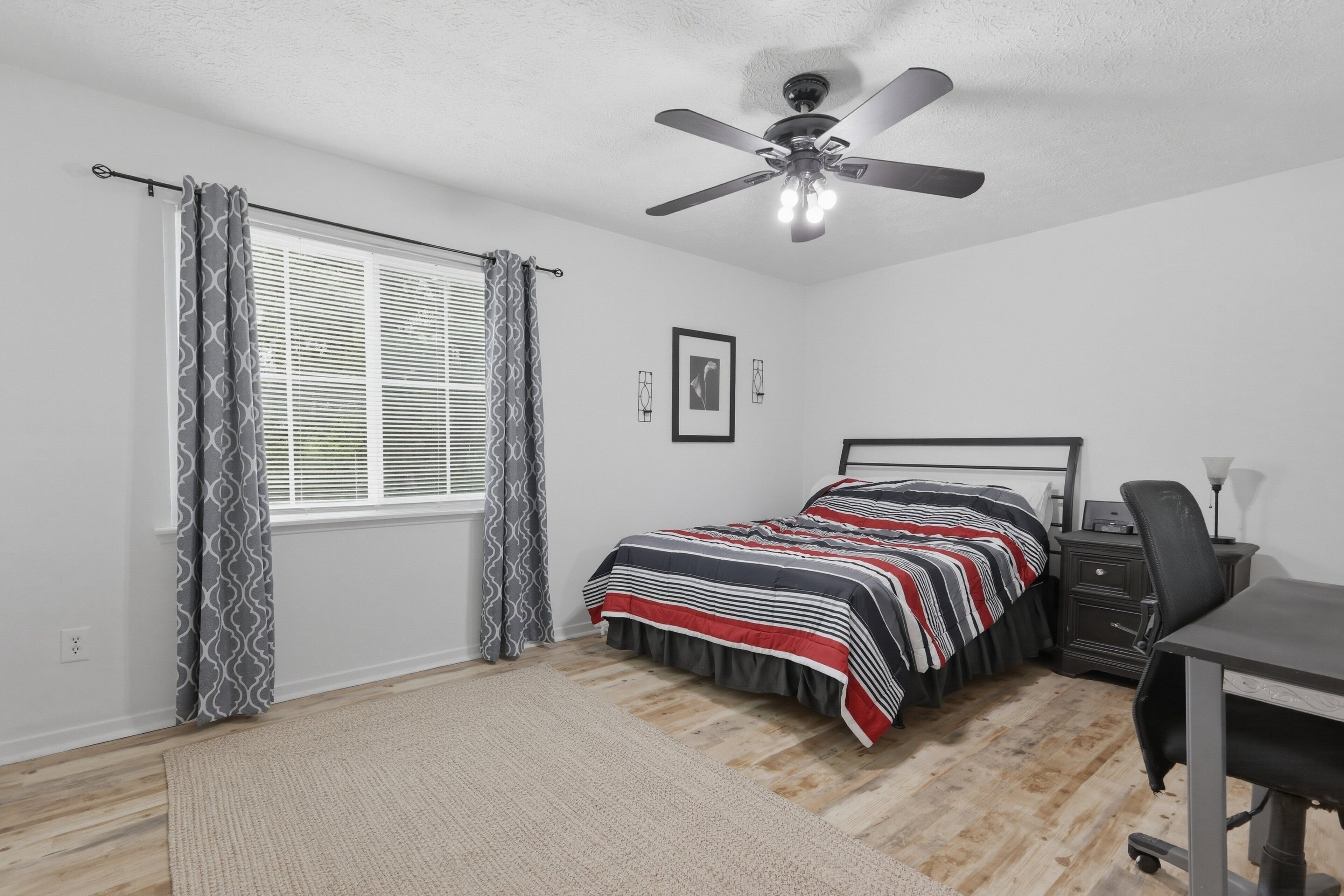 A bedroom with a bed, desk, chair, ceiling fan, and window with curtains.