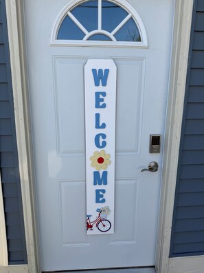Een witte deur met een 'WELCOME' bord, waarop een bloem en een fiets staan.