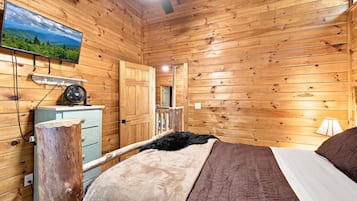 A wooden cabin interior with a bed, a dresser, a television mounted on the wall, and a ceiling fan.