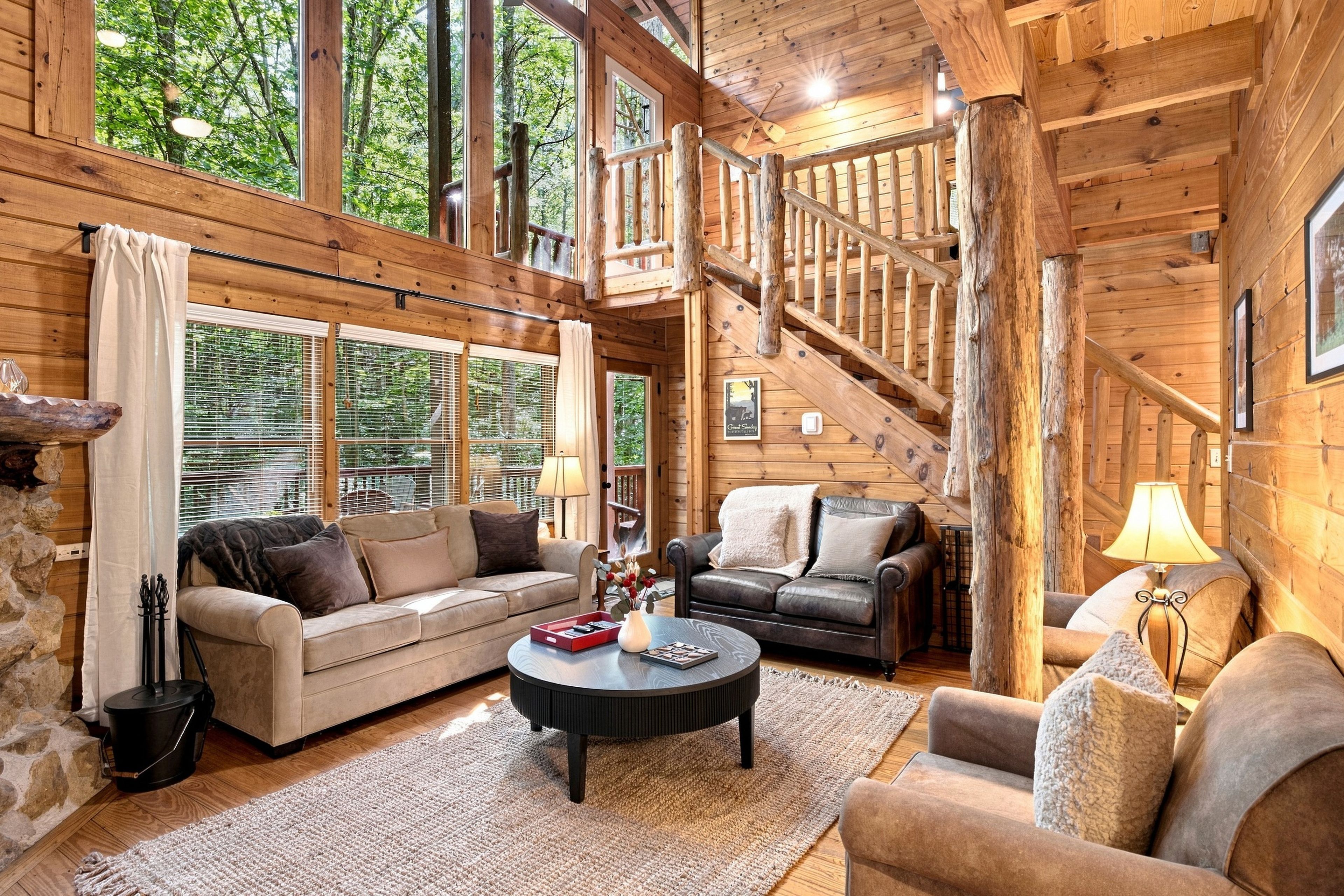 A log cabin interior with a stone fireplace, wooden walls, a staircase, and a cozy seating area with two sofas and a coffee table.