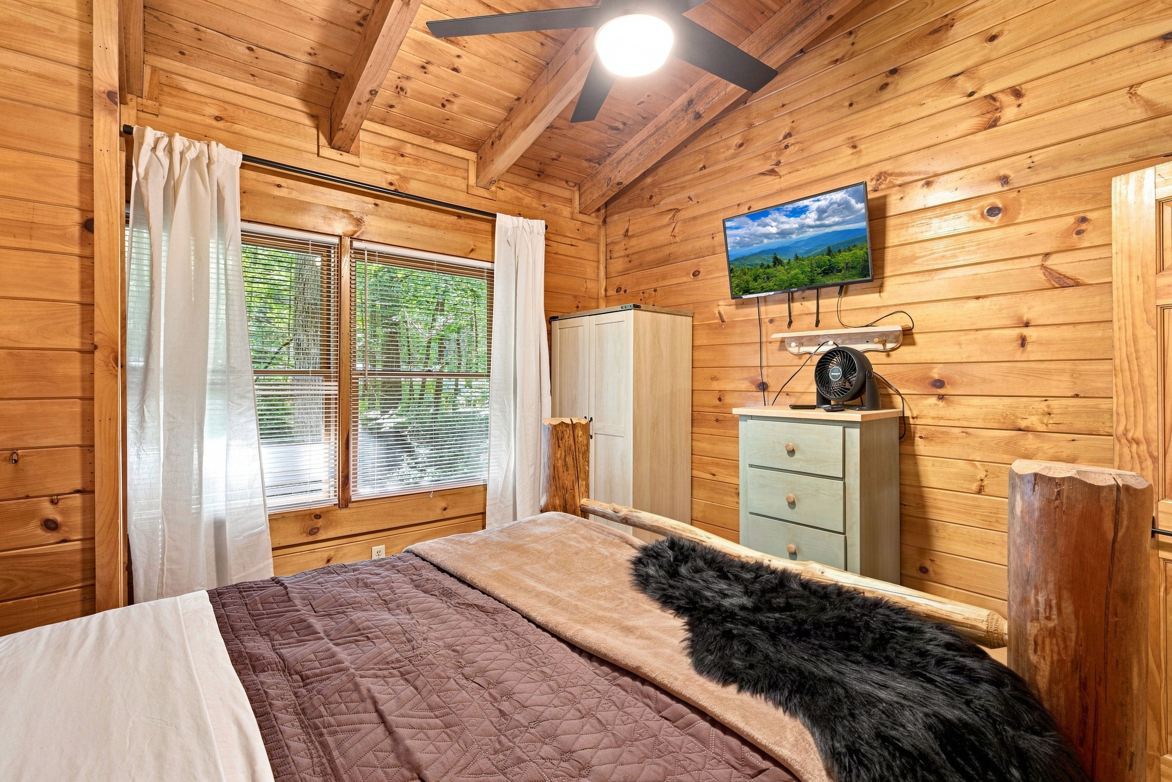 Une chambre dans une cabane en bois, avec un lit, une commode, un téléviseur fixé au mur et une fenêtre avec des rideaux.