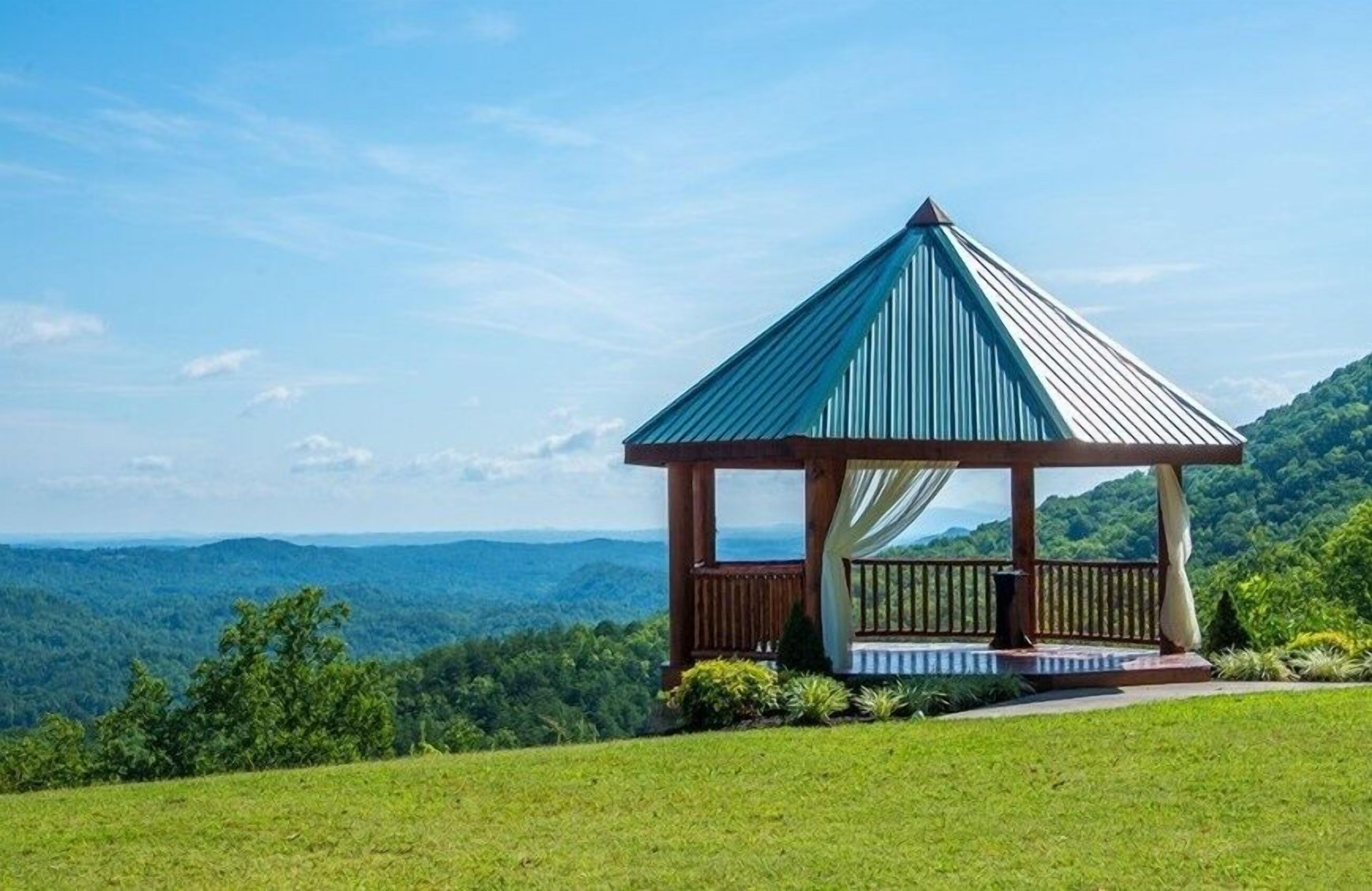 Um gazebo com cobertura metálica e estrutura de madeira, cercado por um gramado e com vista para uma paisagem montanhosa.