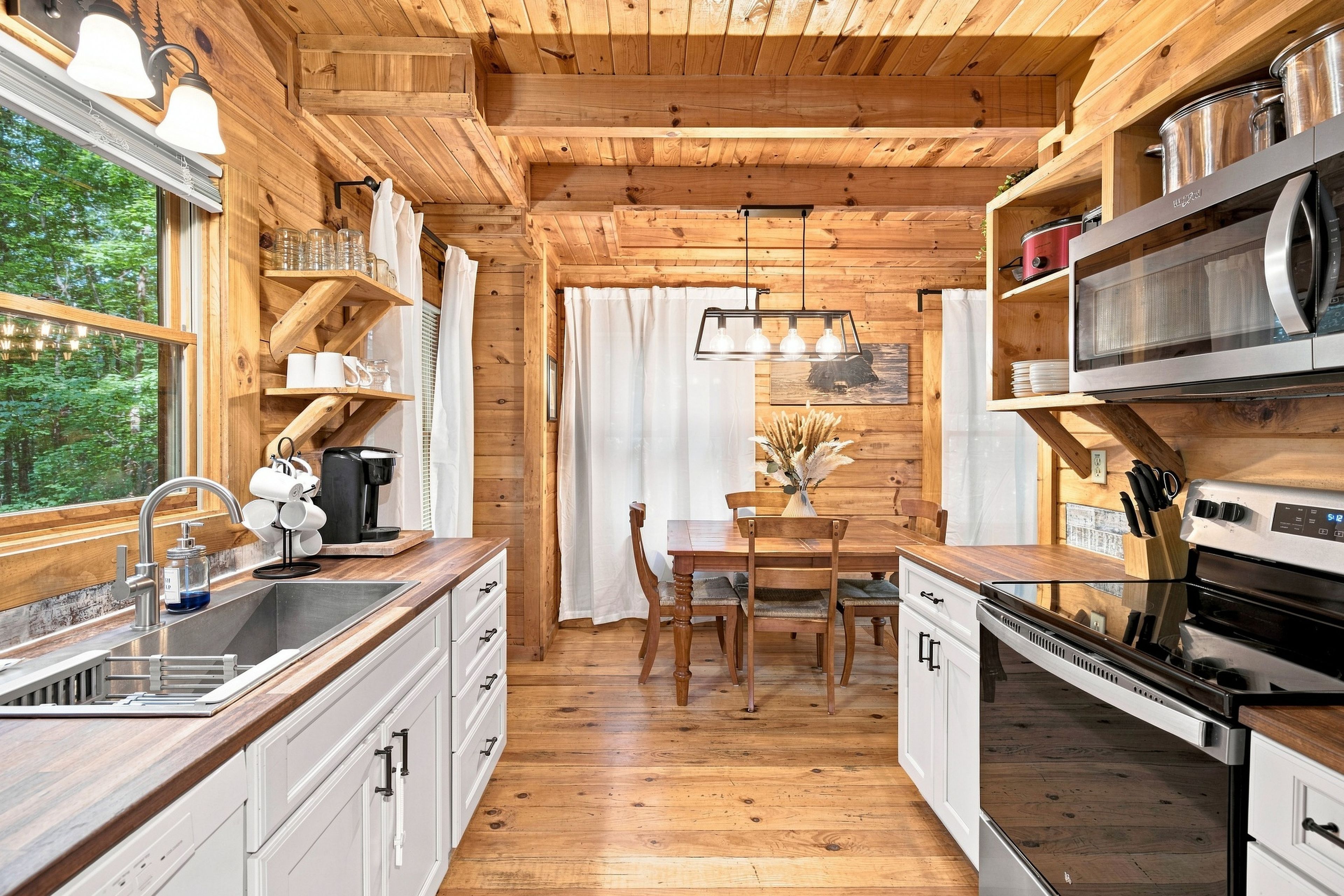 A wooden cabin interior with a kitchen, dining area, and a view of the forest.