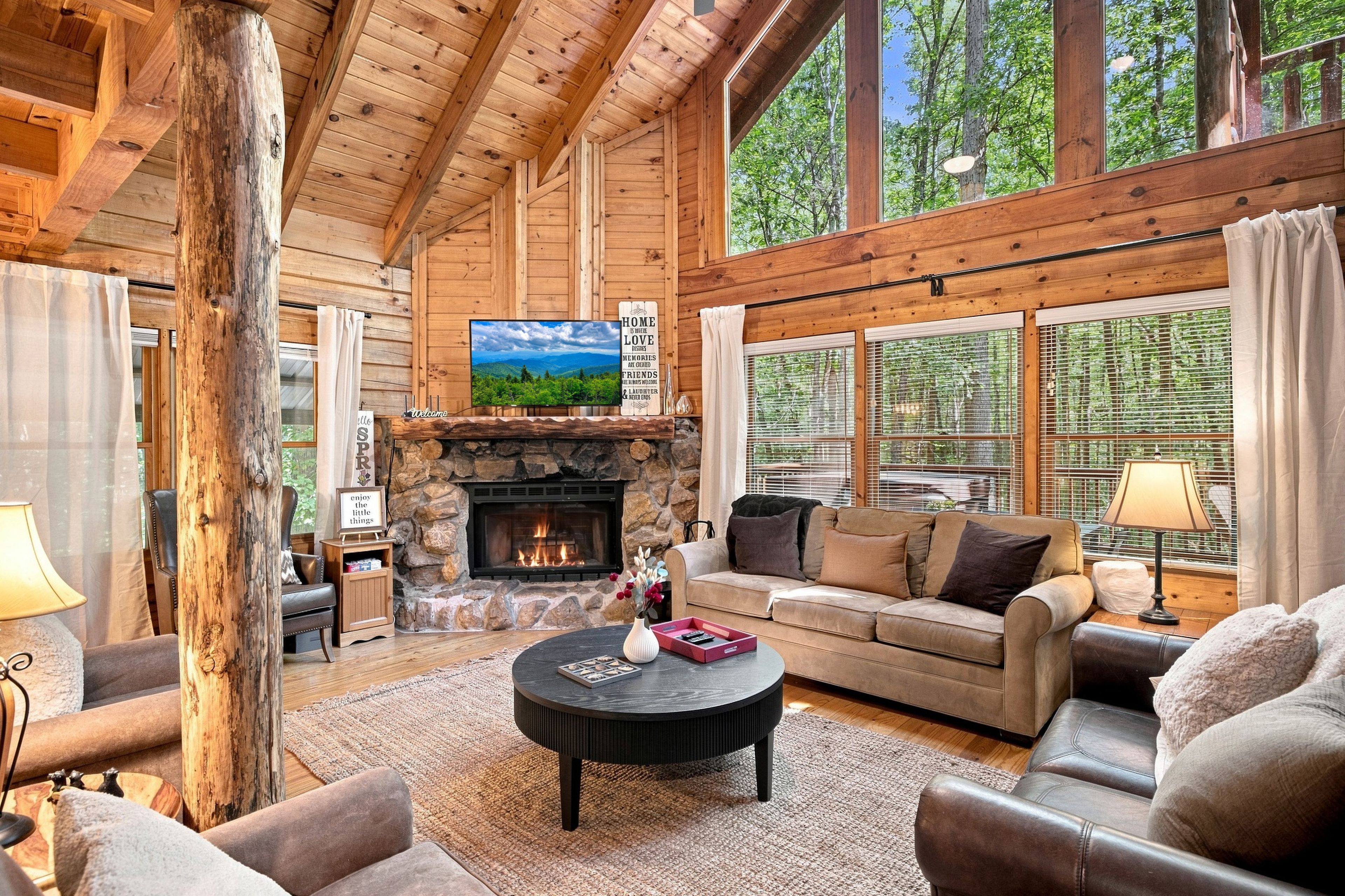 A cozy log cabin interior with a stone fireplace, wooden beams, a comfortable sofa, and large windows.