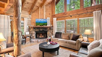 A cozy log cabin interior with a stone fireplace, wooden beams, a comfortable sofa, and large windows.
