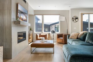 A modern living room with a fireplace, a large window, a sofa, a coffee table, and a wall-mounted TV. - Emerald Escape 3 Bedroom Home (Winter Park)