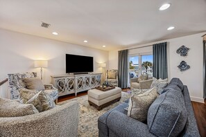 A living room with a flat-screen TV, patterned sofas, a decorative cabinet, and a view through a large window. - 733b Tarpon Avenue 3 Bedroom Apts (Fernandina Beach)