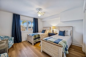 A hotel room with two beds, a ceiling fan, a window with curtains, a bedside table with a lamp, and a small table with a chair. - 733a Tarpon Avenue 3 Bedroom Apts (Fernandina Beach)