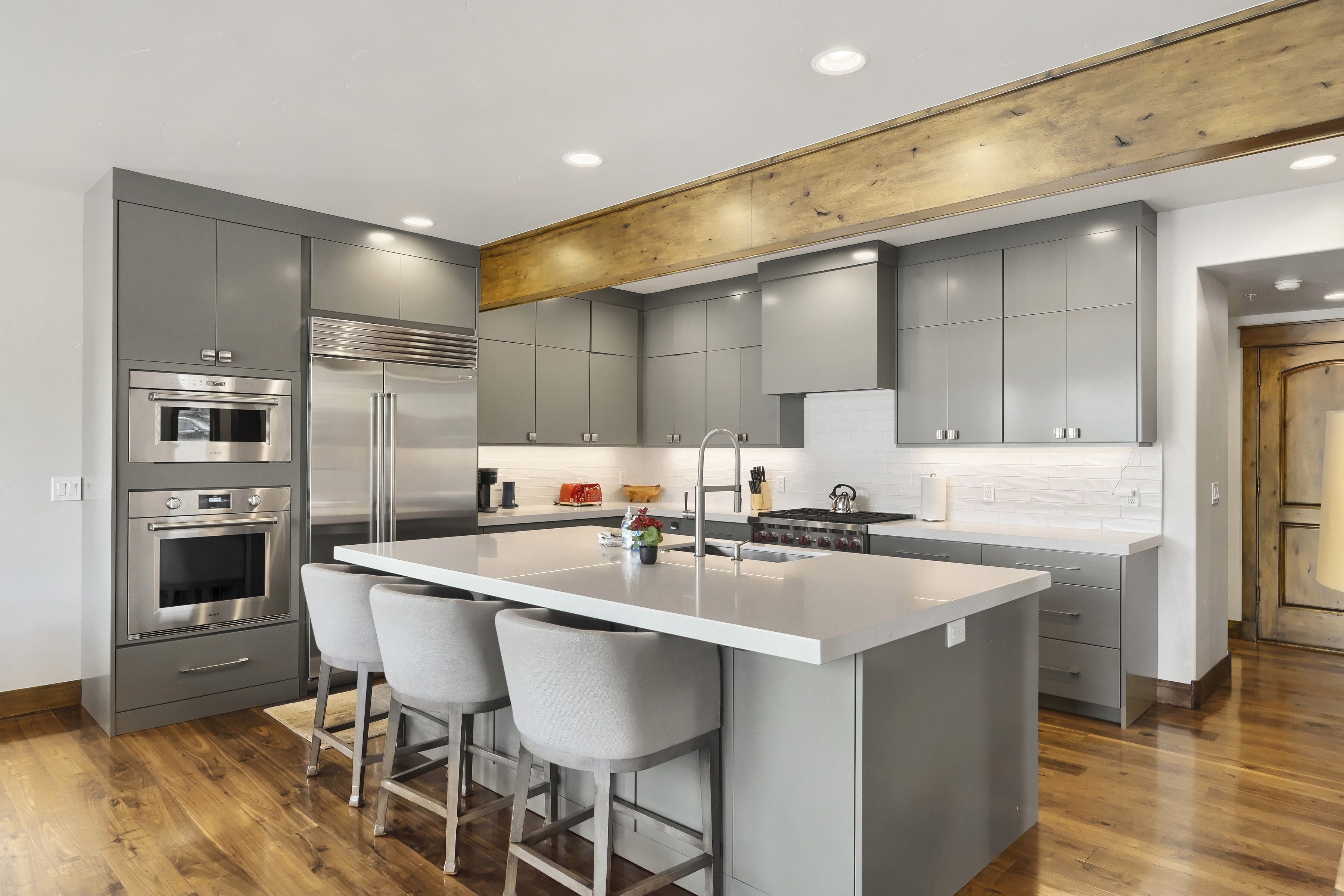 A modern kitchen with grey cabinetry, stainless steel appliances, a central island with bar stools, and a wooden ceiling beam.