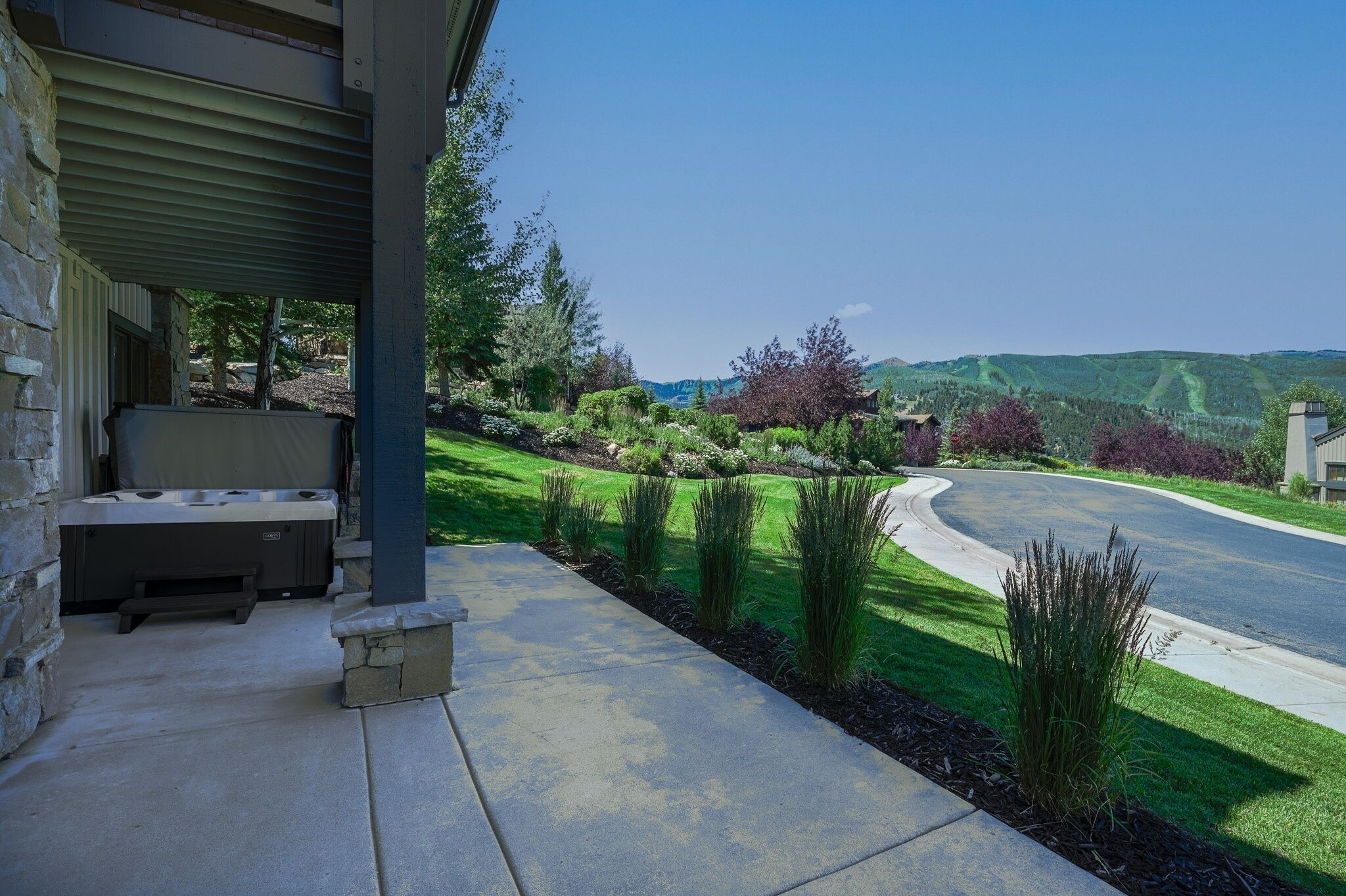 A covered outdoor area with a hot tub, a stone bench, and a view of a landscaped garden and road.