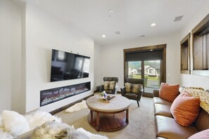 Ein modernes Wohnzimmer mit Flachbildfernseher, Kamin, Ledersofas und einem großen Fenster mit Blick auf ein Haus.