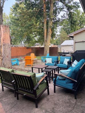 Een terras buiten met zitplaatsen, waaronder stoelen en een bank, omringd door bomen en een houten hek.