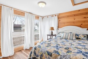 Quarto com cabeceira de madeira, cama com colcha, mesa de cabeceira com abajur e janela com cortinas.