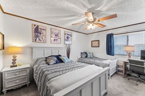 Ein Schlafzimmer mit zwei Betten, einem Deckenventilator, gerahmten Bildern an der Wand, einem Schreibtisch mit Stuhl und einem Fenster mit Vorhängen.
