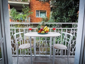 Un balcón con una mesa redonda y dos sillas, un frutero sobre la mesa, y vistas a un edificio de ladrillo y follaje.