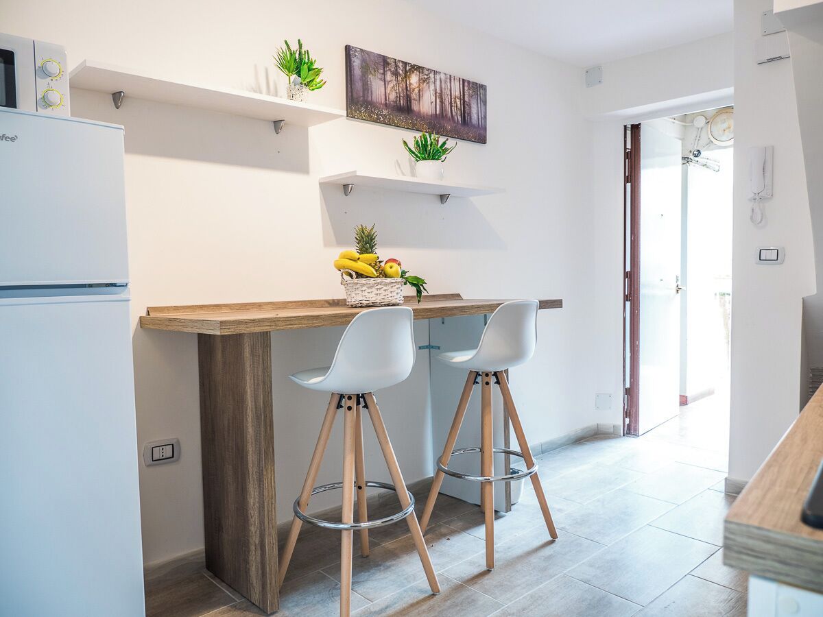 Una cucina moderna con un piano di lavoro in legno, due sgabelli da bar bianchi e un frigorifero. Si vedono piante su mensole sospese e un quadro al muro.