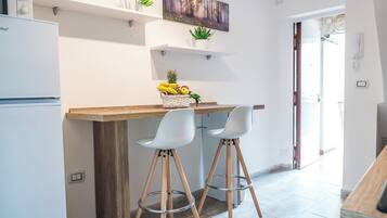 Una cucina moderna con un piano di lavoro in legno, due sgabelli da bar bianchi e un frigorifero. Si vedono piante su mensole sospese e un quadro al muro.