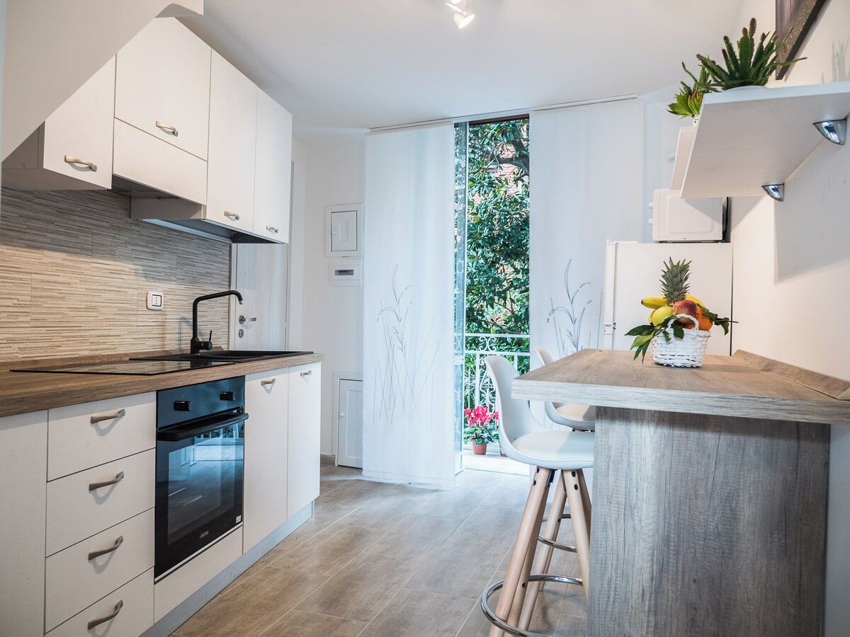 Una cocina moderna con armarios blancos, encimera de madera y horno empotrado. Se aprecia una pequeña isla con una cesta de frutas y una silla.
