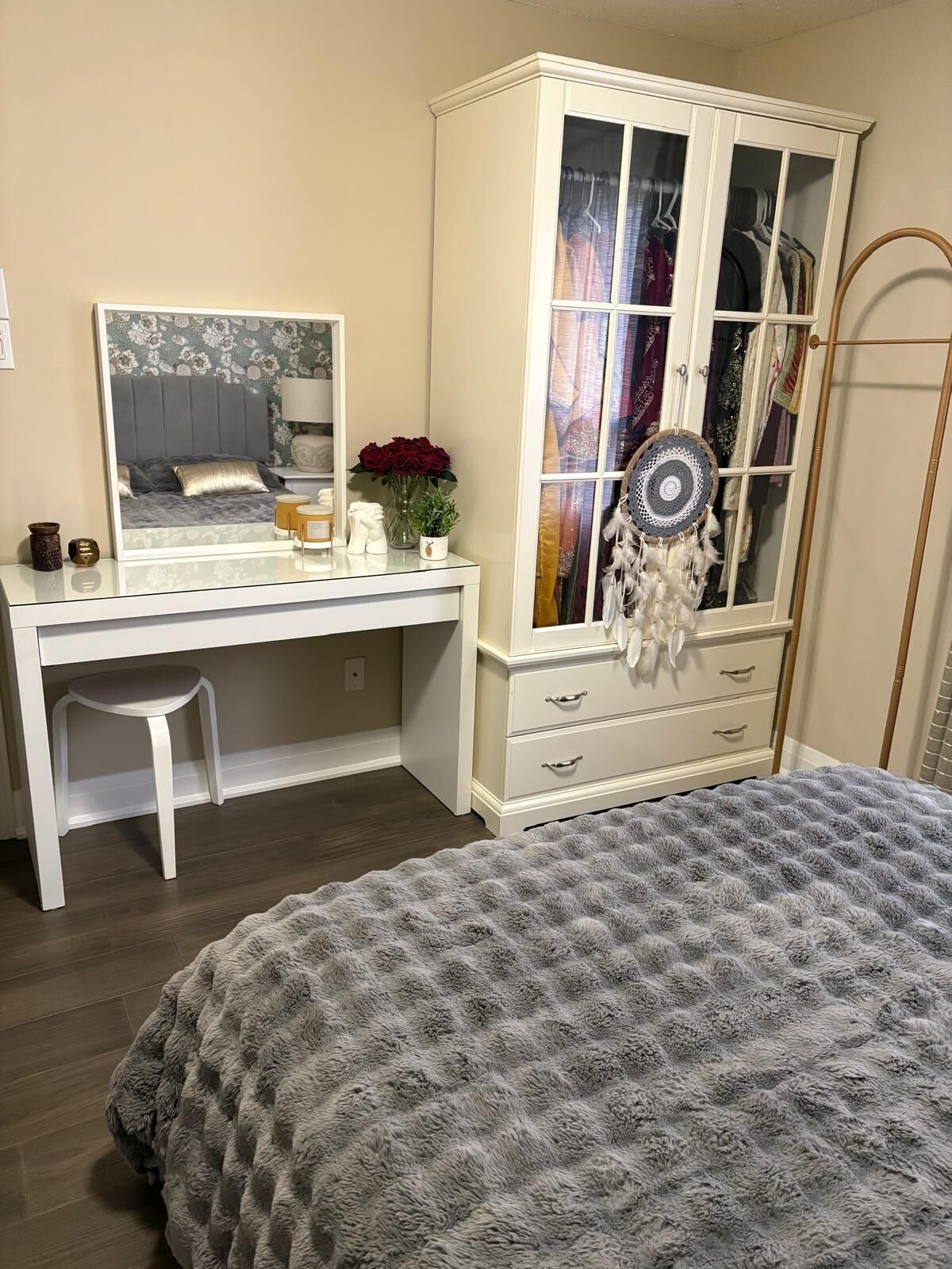 A bedroom with a bed, a white dresser with drawers, a vanity with a mirror, and a wardrobe with glass doors.