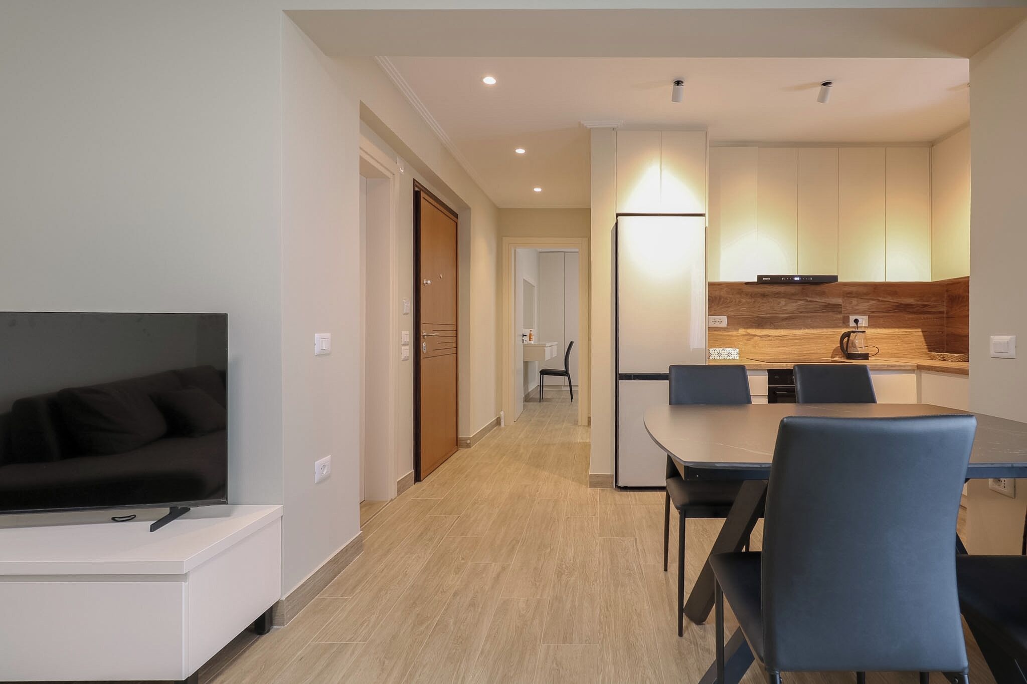 Uma sala de estar moderna com televisão de tela plana, área de jantar com mesa e cadeiras, e cozinha com armários de madeira e cafeteira.