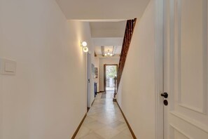 Un couloir avec une porte sur la droite, un escalier sur la gauche et, au fond, une vue sur une autre pièce.