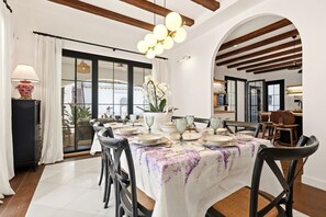 Un coin repas avec une table ronde dressée pour six personnes, entourée de chaises noires, recouverte d’une nappe blanche ornée de motifs floraux.