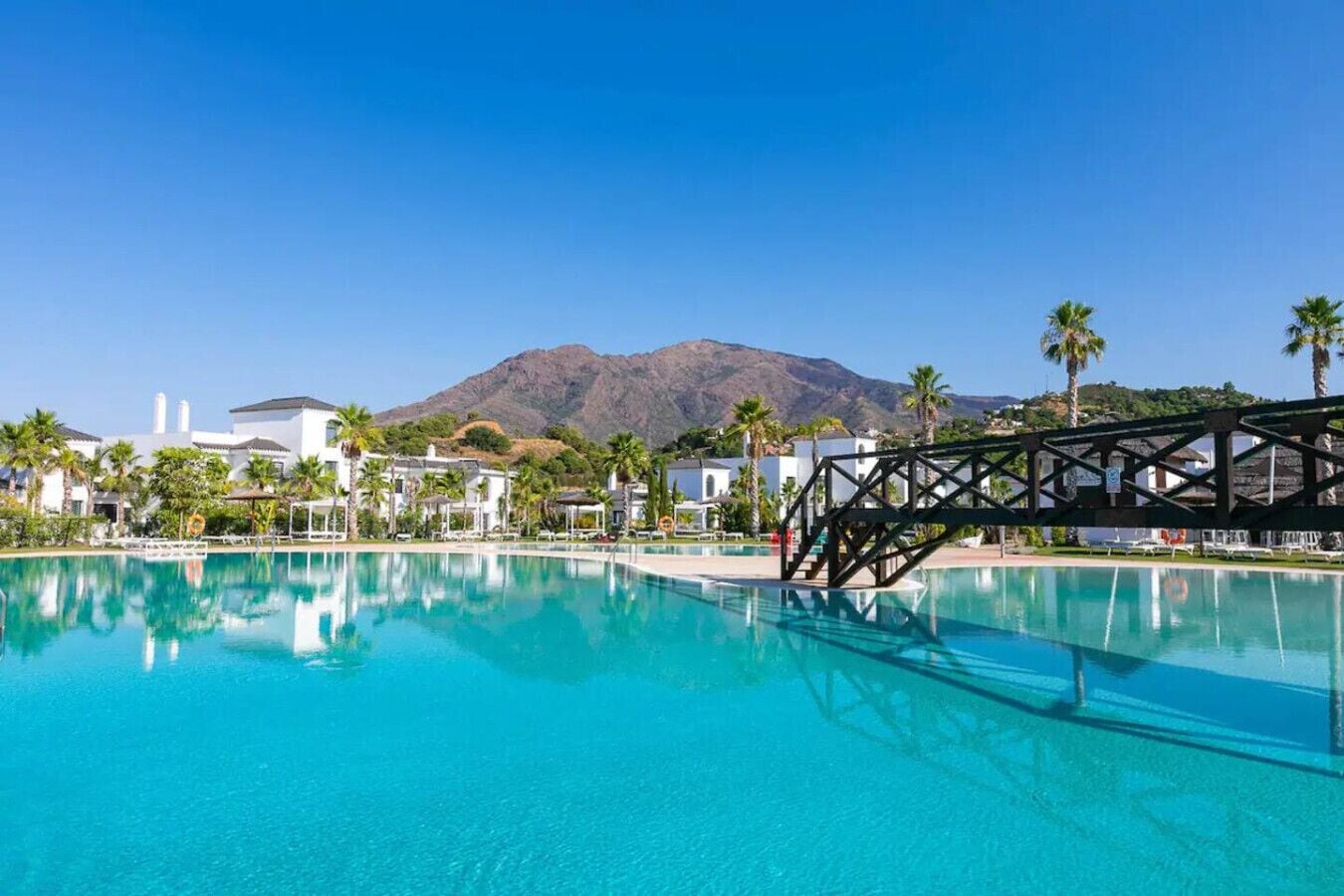 A clear blue pool with a wooden deck structure, surrounded by white buildings and palm trees, with a mountain in the background.