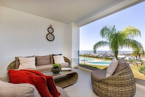 A balcony with a view of a pool and cityscape, furnished with a sofa, armchair, and a red throw blanket. - Estepona Hills Luxury Resort 3-Bedroom Apartment (Estepona)