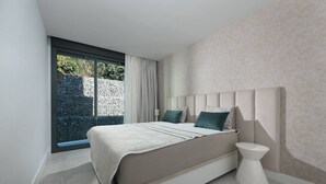 A modern bedroom with a large bed, a bedside table, and a view of a garden through a sliding door. - Sky Penthouse with Private Pool (Artola)