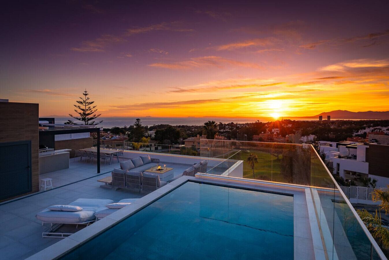 Ein Pool auf dem Dach mit Liegestühlen und Blick auf den Sonnenuntergang über einer Stadtlandschaft.
