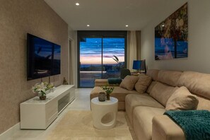 A modern living room with a flat-screen TV, a sofa, a coffee table, and a view of the sunset through a large window. - Sky Penthouse with Private Pool (Artola)