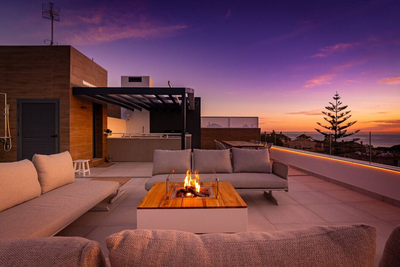 A rooftop terrace with a sofa set, a coffee table with a fire feature, and a view of the sunset.