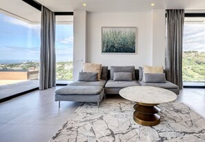A modern living room with a sectional sofa, a round coffee table, and a large window with curtains. - Duplex Penthouse · Sea & Sky · Artola Alta (Marbella)