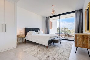 Ein modernes Schlafzimmer mit einem großen Bett, einem Nachttisch, einem Sessel, einem Schreibtisch und Blick ins Freie.