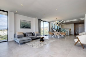 A modern living room with a sectional sofa, dining table, and chairs, a large window with a city view, and a kitchen area in the background. - Duplex Penthouse · Sea & Sky · Artola Alta (Marbella)