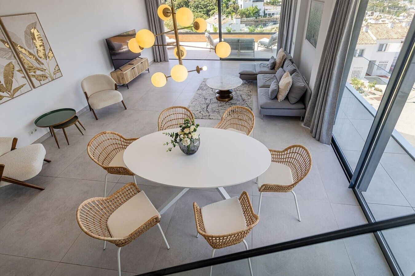 A modern living room with a round table, wicker chairs, a sofa, and a large window offering a city view.