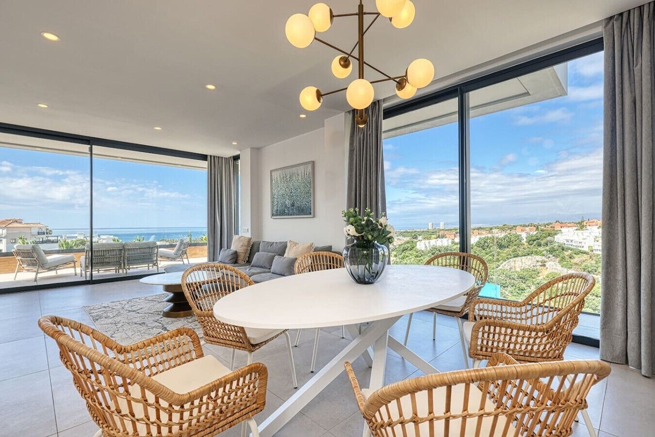 A spacious room with a round dining table surrounded by wicker chairs, large windows offering a view of the sea, and a modern chandelier.