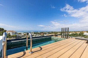 Ein Pool auf dem Dach mit einer Holzterrasse und Metallgeländern, mit Blick auf eine Küstenstadt.