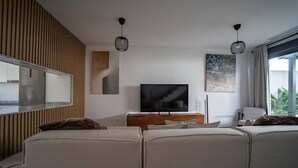 A modern living room with a flat-screen TV, a wooden accent wall, a large window with curtains, and a sofa. - Charming Townhouse in Cancelada Village (Cancelada)