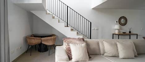 Een moderne woonkamer met een bank, een eettafel met stoelen, een trap en een bijzettafeltje met een lamp en decoratieve objecten.