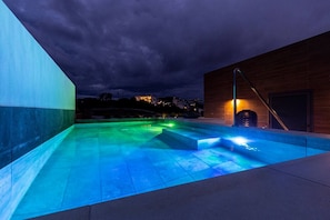 Ein Außenpool bei Nacht mit blauer Beleuchtung, ein Holzgebäude und im Hintergrund ein Blick auf eine Stadtlandschaft.
