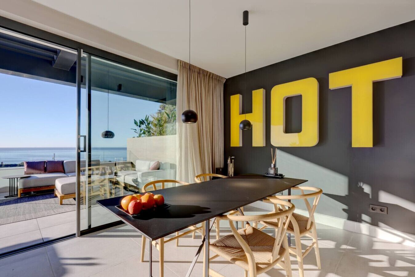 A modern dining area with a glass wall overlooking the sea, a table with fruits, and a bold 'HOT' sign on the wall.