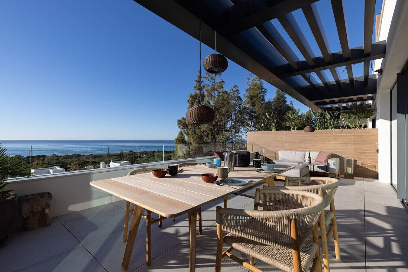 An outdoor patio with a table and chairs, a hanging light fixture, and a view of the ocean.