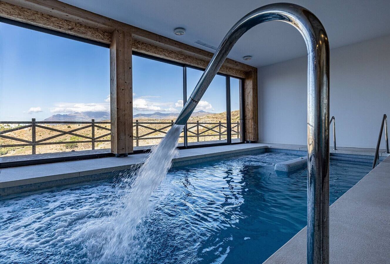 An indoor swimming pool with a waterfall feature, large windows, and a view of a mountainous landscape.