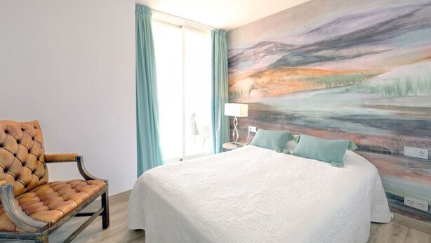 A hotel room with a large bed, a tufted armchair, a painting on the wall, and a window with curtains. - Marbella La Ola: 18th Floor (Marbella)