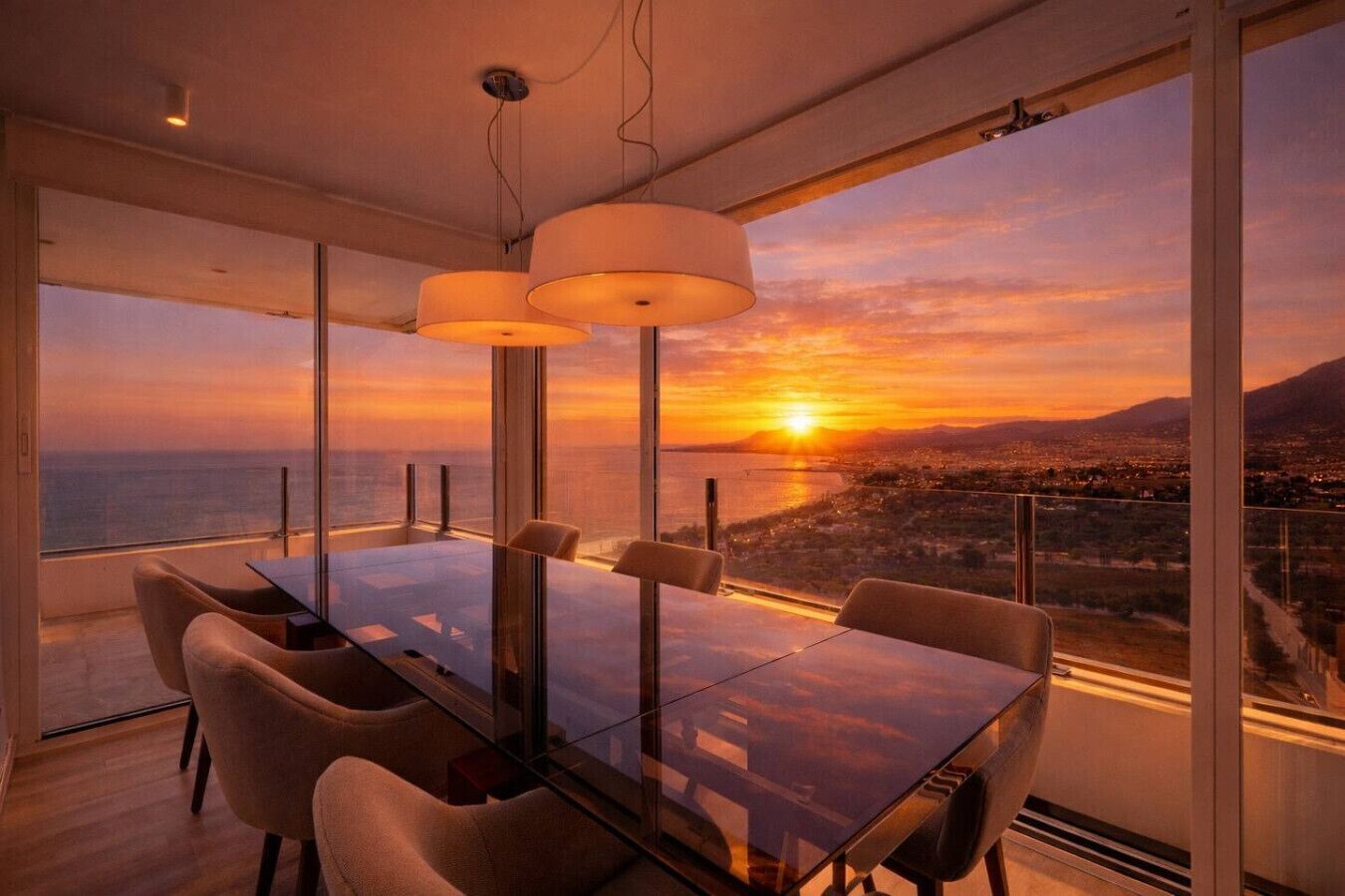 Comedor con mesa y sillas de vidrio, ventanas amplias y vista al atardecer.