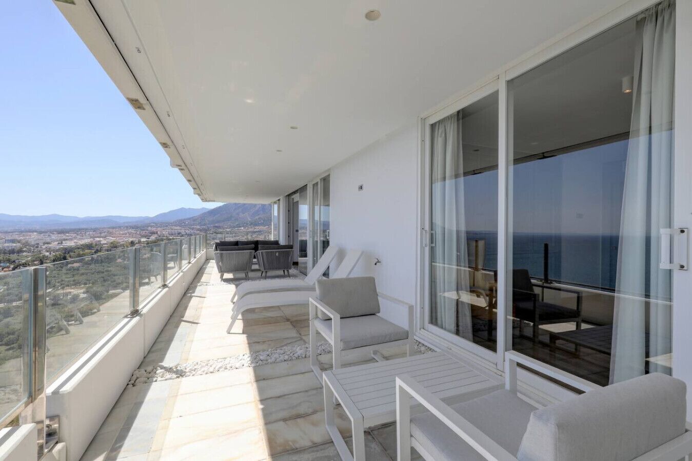 A balcony with a view of the sea and mountains, furnished with white chairs and a table.