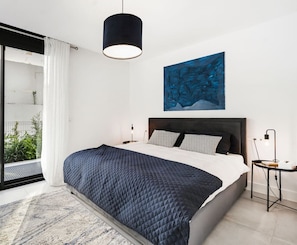 A modern bedroom with a large bed, a black pendant light, a nightstand, and a view of greenery through a sliding door. - Luxury Apartment with Ocean View (Marbella)