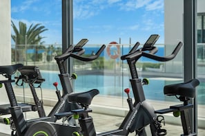 Varias bicicletas estáticas con pantallas digitales, ubicadas cerca de una ventana con vista a una piscina y palmeras.